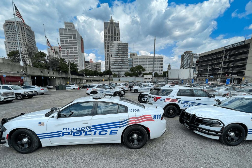 Detroit Police vehicles are parked on a fenced lot with the downtown skyline on the background