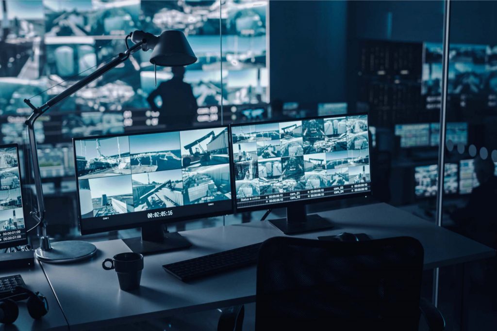 Two digital computer screens with surveillance CCTV video in a port monitoring center with multiple cameras on a large digital screen.