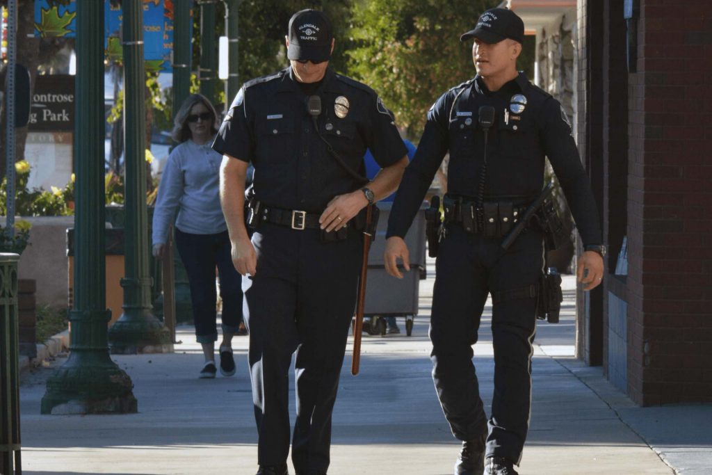 Two policemen stroll toward the camera along a quiet tree lined sidewalk